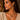 A close-up of a woman wearing a gold chain necklace with a triangular labradorite pendant and a matching gold hoop earring, paired with a beige ribbed knit top.