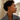 A close-up side profile of a woman with short curly black hair wearing a pair of gold huggie hoop earrings with oval bezel-set clear stones