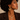 A close-up of a woman wearing a gold necklace with a circular medallion pendant and a large twisted hoop earring, paired with a dark brown blazer against a warm beige background.