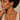 A close-up of a woman wearing a gold chain necklace with teardrop charms and matching gold hoop earrings with small clear stones, against a warm beige background.